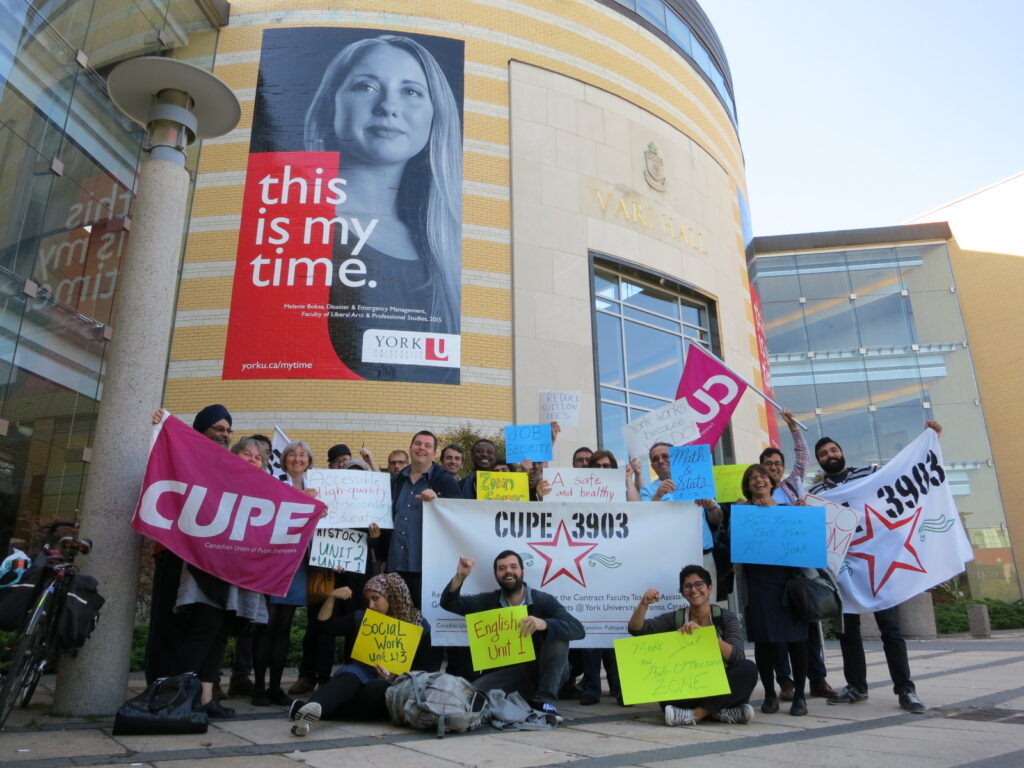 CUPE 3903 members and staff posing in front of Vari Hall at York University with various flags, signs, and banners. Many of the signs indicate the member's unit and department. They are cheering enthusiastically. There is a poster visible on Vari Hall with an image of someone posing and text that reads "this is my time".