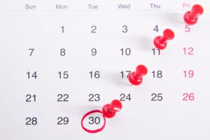 A month-long calendar with a diagnoal row of red thumbtacks placed in it and the date "30" circled in red.