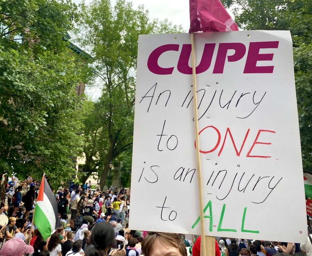 A CUPE sign and flag in the foreground of a crowded rally at the University of Toronto, with trees and a Palestine flag visible in the background. The sign reads: “CUPE - An injury to ONE is an injury to ALL”.