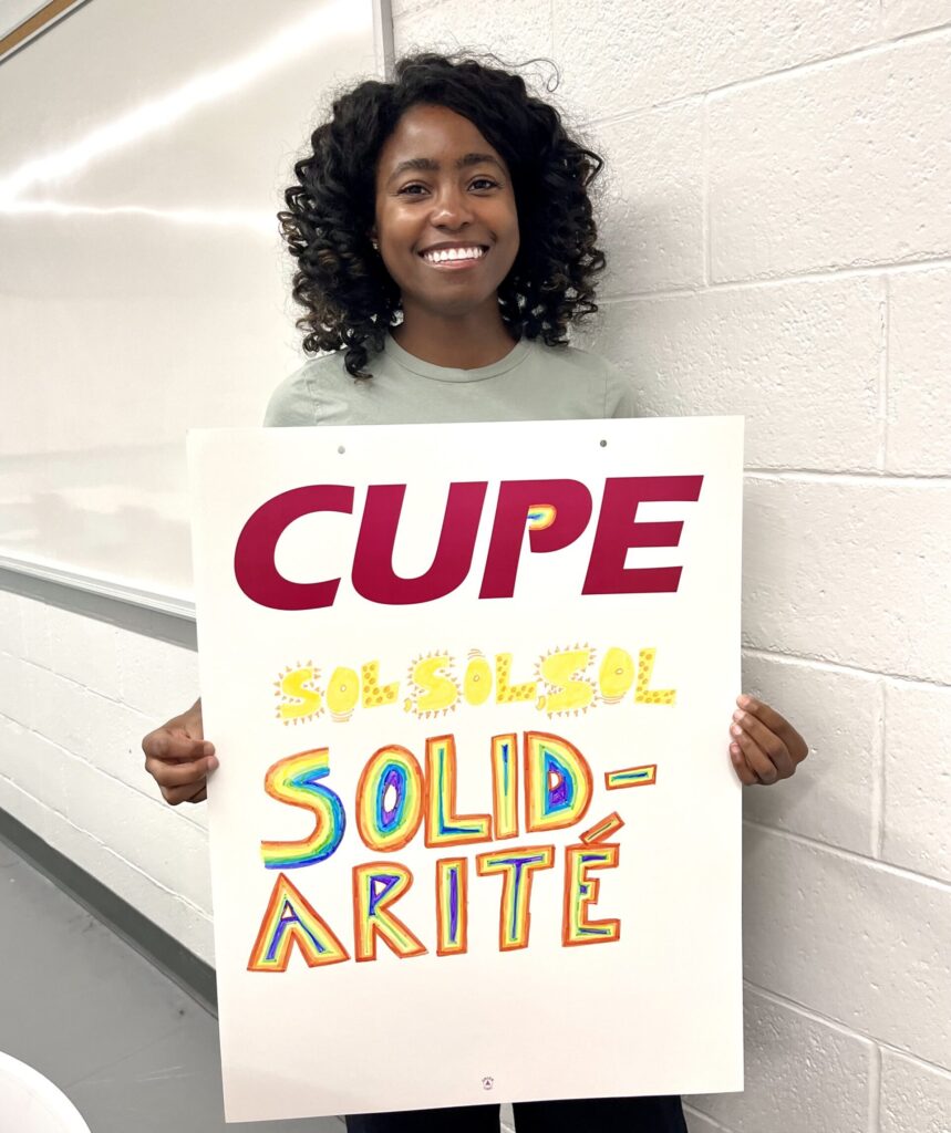 A member holding a hand decorated sign that reads “Sol Sol Sol Solidarité” with rainbow letters.