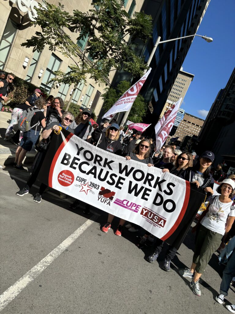 An image of CUPE 3903 and other union members at the Labour Day Parade, holding a banner with the logos of various unions on campus. The text of the banner reads "York Works Because We Do".