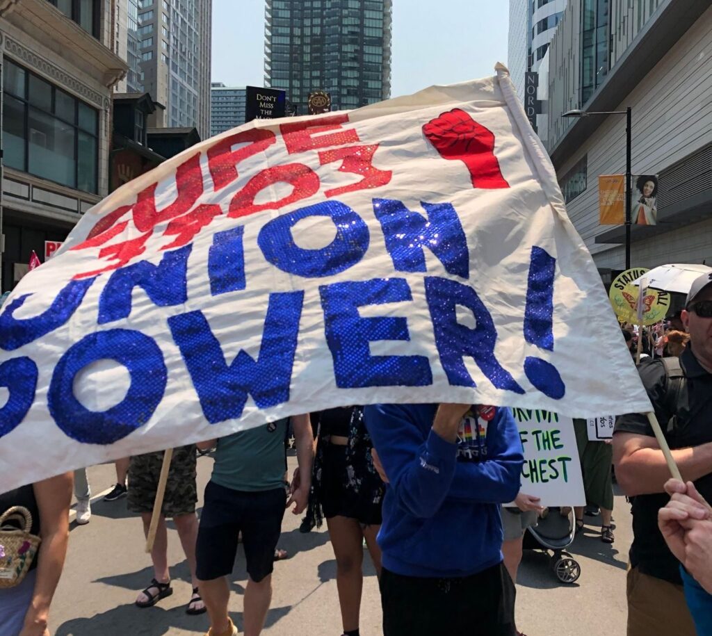 CUPE 3903 members at the Ontario Federation of Labour 'Enough is Enough' rally, with a historic sequined homemade flag from the 2001 strike in the foreground. It reads "CUPE 3903 Union Power!" in red and blue lettering, with a fist symbol.