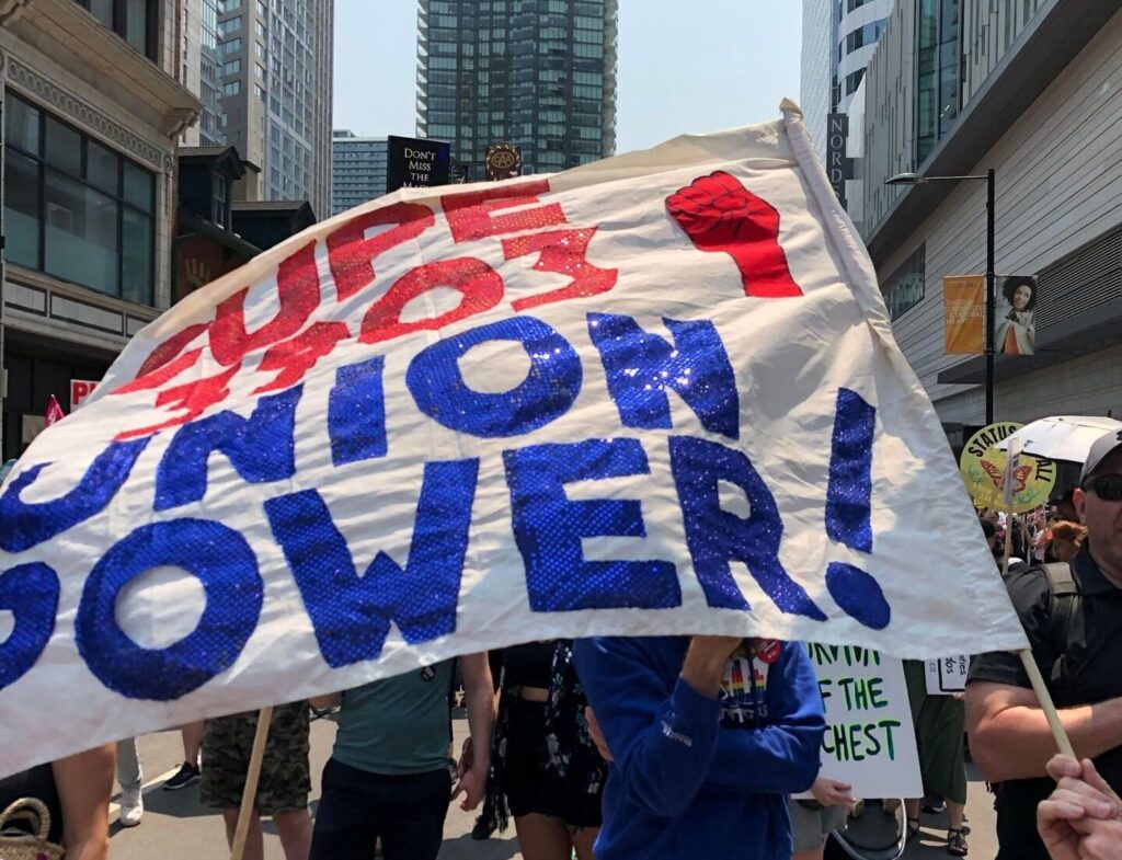 CUPE 3903 members at the Ontario Federation of Labour 'Enough is Enough' rally, with a historic sequined homemade flag from the 2001 strike in the foreground. It reads "CUPE 3903 Union Power!" in red and blue lettering, with a fist symbol.