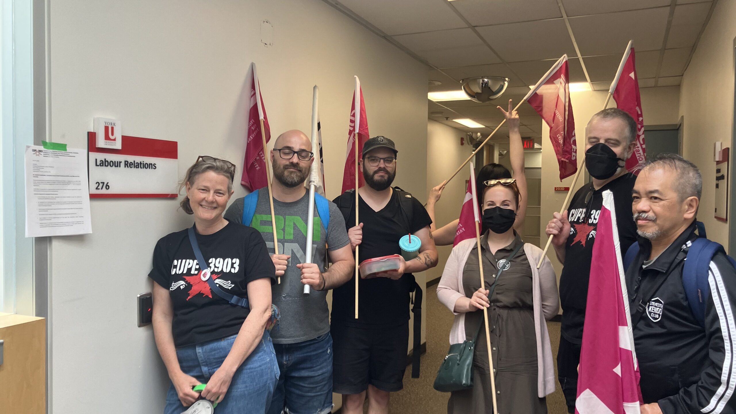 Members holding flags stand in front of the Labour Relations Office, with the Notice to Bargain taped to the doorframe. 