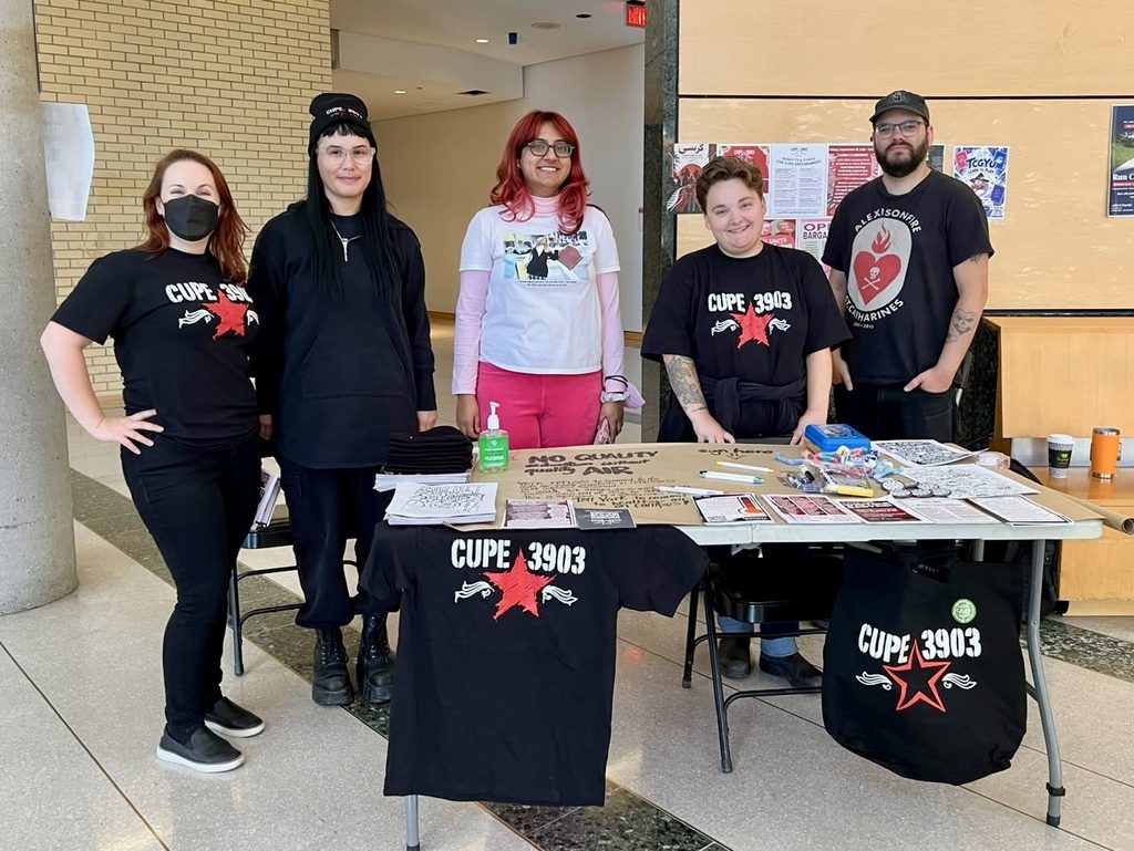 Five CUPE 3903 members standing behind a table in Vari Hall. The table is adorned with union flyers and apparel.