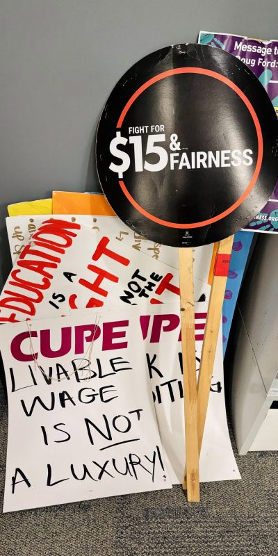 A pile of various picket signs, including ones that read "$15 and fairness" and "CUPE - Living wage is NOT a luxury".