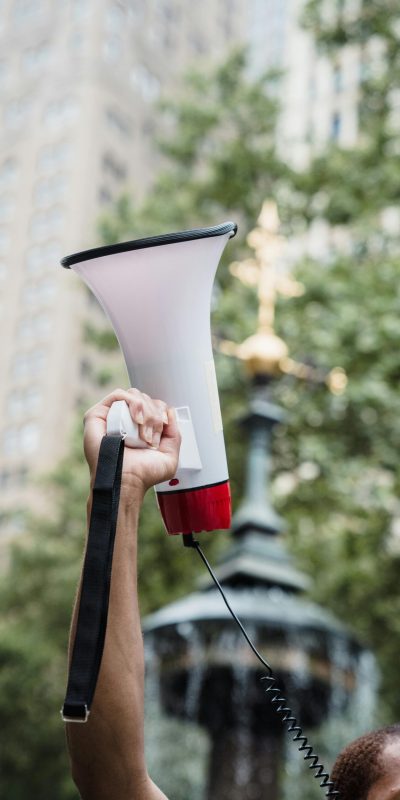 In the foreground, someone's arm is visible, holding a white megaphone up in the air. In the background, city architecture and greenery are visible.